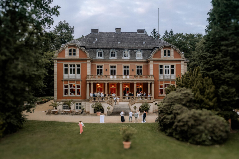 Frontalansicht von Schloss Eldingen mit einer Veranstaltung auf der Terrasse und Gästen im Garten.