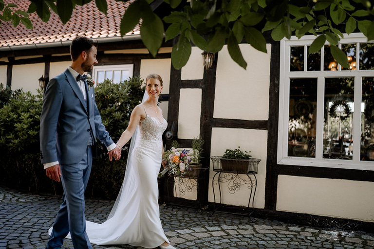 Braut und Bräutigam spazieren Hand in Hand durch den Fachwerkhof der Moormühle