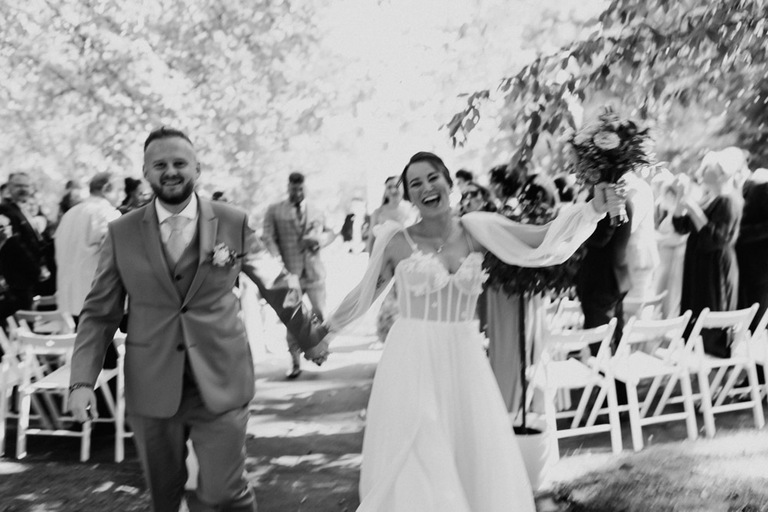 Die Braut und der Bräutigam laufen nach der Trauung strahlend durch den Waldgarten.