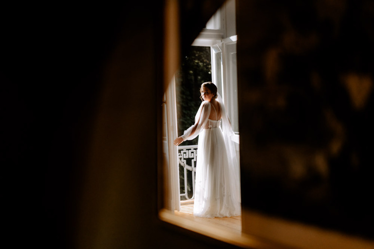 Braut in einem eleganten Kleid steht vor einem großen Fenster im Schloss Eldingen.