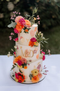 Dreistöckige Hochzeitstorte mit bunten Blumen und goldenen Details, verziert mit den Initialen M&D