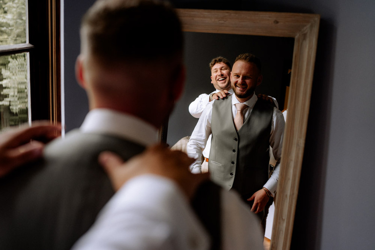 Der Bräutigam lacht vor einem Spiegel, während ein Freund ihm die Schultern klopft.