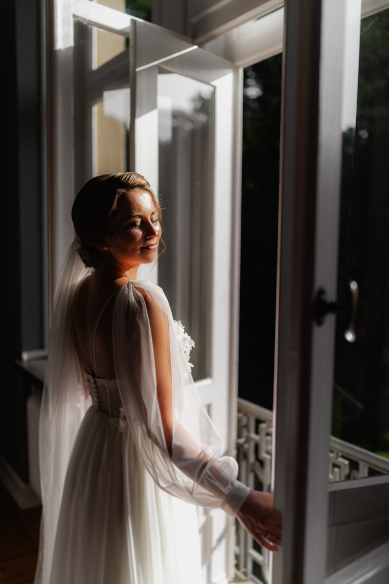 Die Braut lächelt sanft, während sie an einem geöffneten Fenster von Schloss Eldingen steht.